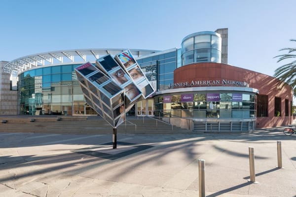 The glass and steel facade of the Japanese American National Museum in Los Angeles, gleaming in sunlight as visitors gather outside in the Little Tokyo district.